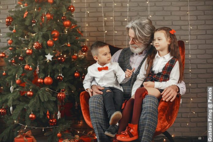 An elderly man sitting on a chair with his grandchildren beside a Christmas tree