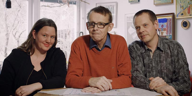 Hans Rosling seated between his son Ola Rosling and daughter-in-law Anna Rosling Rönnlund at a table with papers.